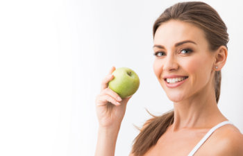 A smiling woman with brown hair in a white top holds a green apple near her face against a plain white background.