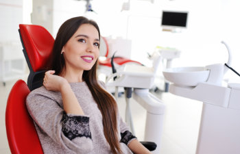 A woman with long hair is sitting in a red dental chair smiling. Dental equipment and a monitor are visible in the background.